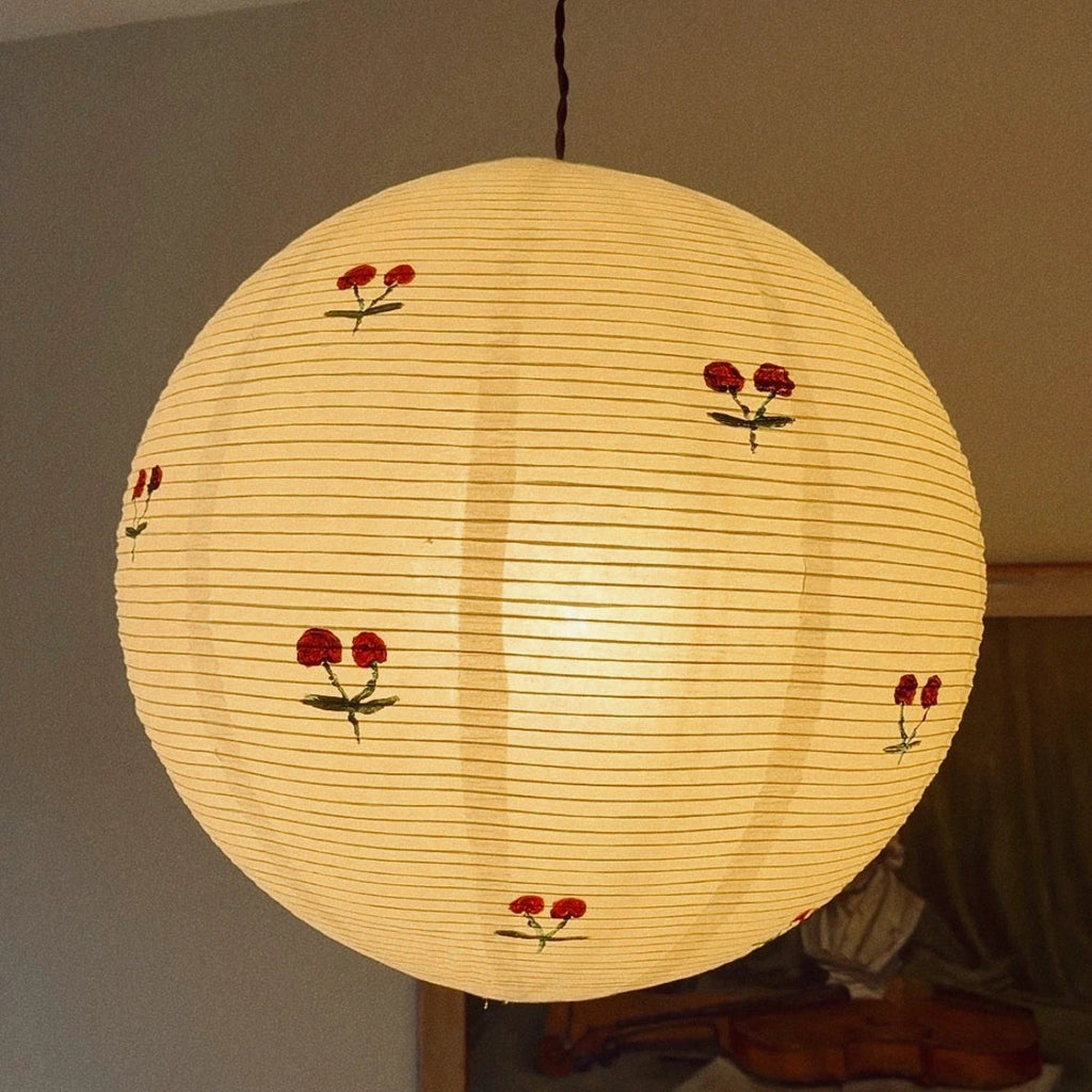 A spherical paper lantern pendant light with a warm yellow glow, decorated with small red cherry blossoms and green stems, hanging in a room.
