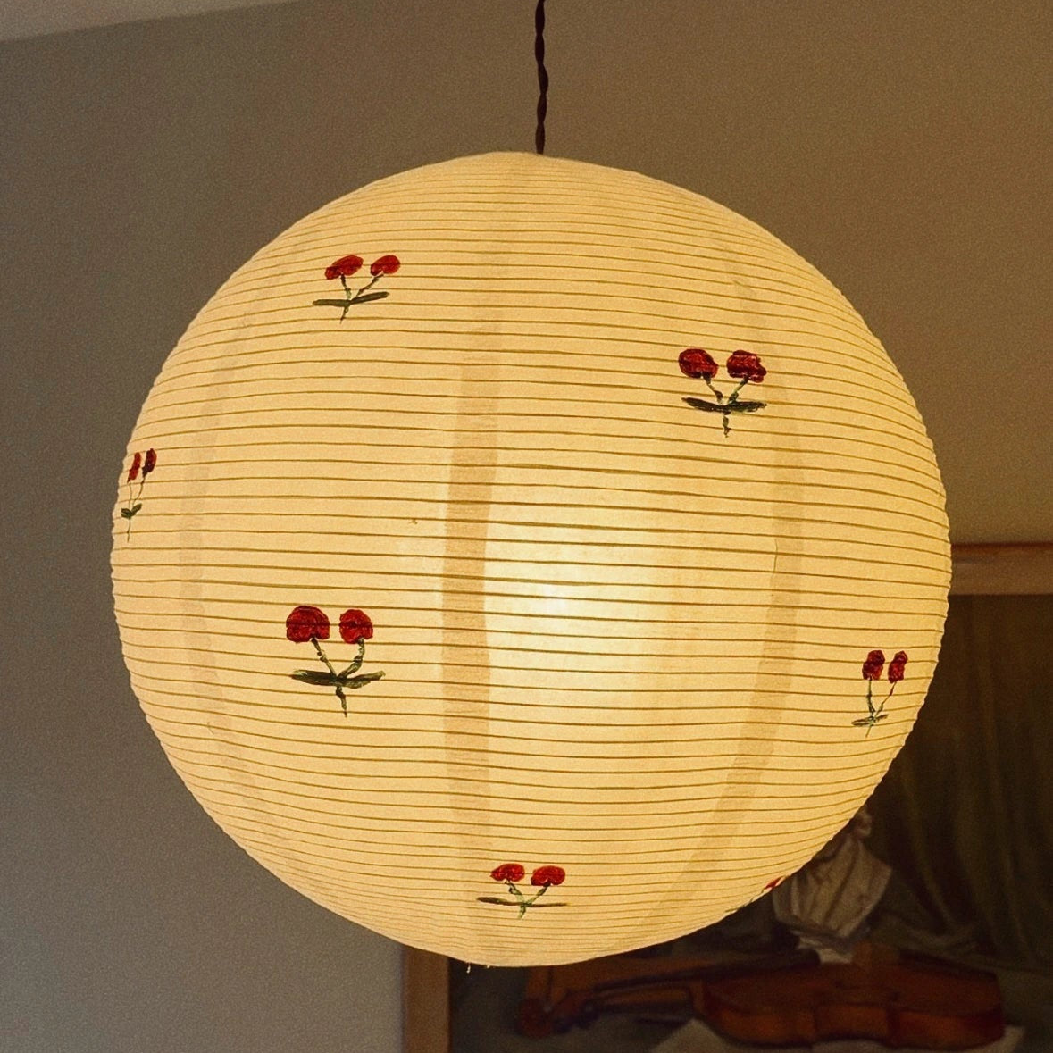 A spherical paper lantern pendant light with a warm yellow glow, decorated with small red cherry blossoms and green stems, hanging in a room.